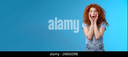 Amazed gasping shocked wondered redhead attractive curly girl popping eyes stare camera drop jaw concerned astonished touch cheeks empathy amazement Stock Photo