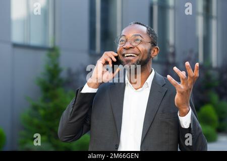 Cheerful and satisfied african american boss outside office building smiling and talking to colleagues on phone, man in business suit walking on lunch break. Stock Photo