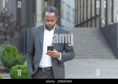 Serious and successful African American boss businessman outside office building using smartphone, man in business suit reading news online thinking and browsing internet pages. Stock Photo