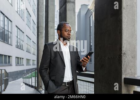 Serious and successful African American boss businessman outside office building using smartphone, man in business suit reading news online thinking and browsing internet pages. Stock Photo