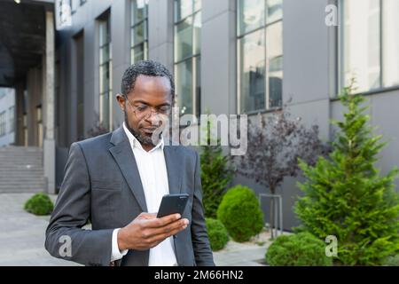 Serious and successful African American boss businessman outside office building using smartphone, man in business suit reading news online thinking and browsing internet pages. Stock Photo