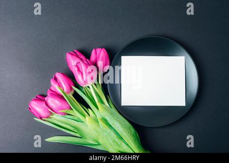 Black plate with blank card and purple tulips on black background. Top view, flat lay, mockup. Stock Photo