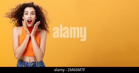 Waist-up shot of charmed enthusiastic attractive female with curly hairstyle in cropped top holding palms on cheeks and smiling astonished and Stock Photo