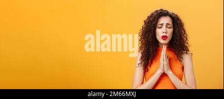 Nervous and concerned woman with curly hairstyle feeling hopeful praying with closed eyes and frowned eyebrows holding hands in pray near chest Stock Photo