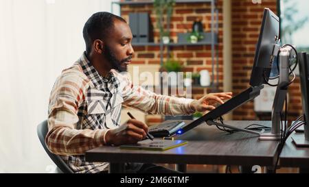African american expert smiling and working with graphic tablet, using stylus pen to develop new project interface. Happy male worker creating artistic artificial intelligence content on computer. Stock Photo