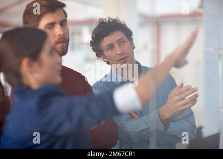 Collaborating on a creative project. A group of creative professionals working together at a glass wall with designs on it. Stock Photo