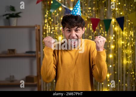 Joyful arabic man in sweater and holiday cap, clenching his fists with happiness, celebrating his birthday, having good mood. Satisfied male posing at Stock Photo