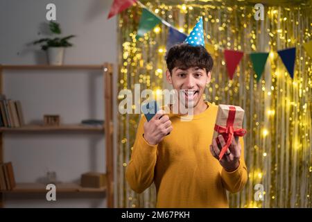 Portrait of arabic guy in festive hat overjoyed cheerful cheery glad excited young man using cell reading great news holding gift box. Home birthday Stock Photo