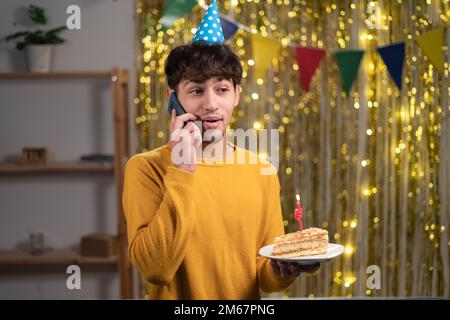 Young arab man celebrating birthday at home talking on cell phone holding birthday cake in hands Stock Photo