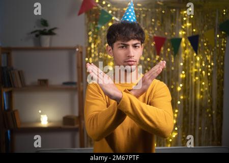 Serious young arab man standing in party hat arms crossed in front of him showing stop sign, celebrating birthday at home, emotions and gestures Stock Photo