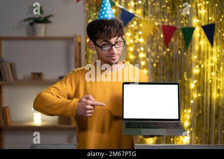 Joyful arabian or indian guy in party hat, freelancer or student, holding an open laptop with empty mock-up space and points finger on it, celebrate Stock Photo