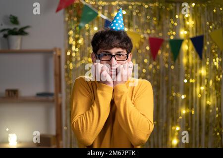 Young arab man in a party hat scared afraid, holding hands near his face, body language and emotions, guy celebrating birthday at home in a bad mood Stock Photo
