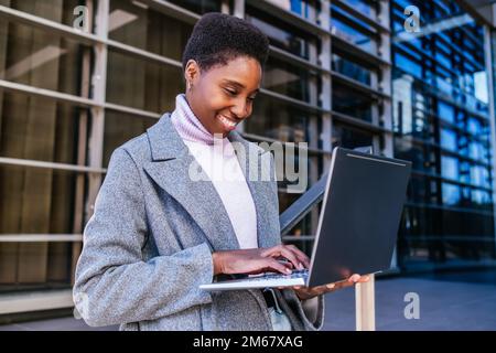 Optimistic young black female in stylish coat and sweater smiling and browsing data on netbook. She is leaning on railing and working on remote project outside contemporary building on city street Stock Photo
