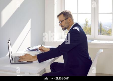 Mature man accountant or auditor working on laptop in office sitting at table, side view. Stock Photo
