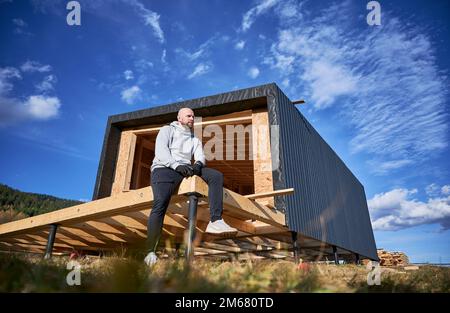 Male carpenter building wooden frame house on pile foundation. Bald man sitting on terrace on construction site, inspecting quality of work. Stock Photo
