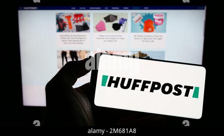 Person holding mobile phone with logo of American news company HuffPost on screen in front of business web page. Focus on phone display. Stock Photo