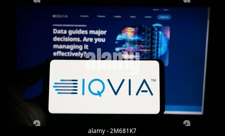 Person holding mobile phone with logo of US health technology company IQVIA Holdings Inc. on screen in front of web page. Focus on phone display. Stock Photo