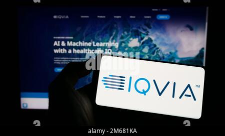 Person holding cellphone with logo of American health technology company IQVIA Holdings Inc. on screen in front of webpage. Focus on phone display. Stock Photo