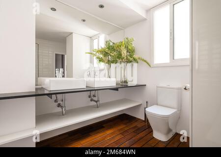 Two square sinks on black marble countertop in spacious bathroom with window to street. Large mirror reflects back of room with white ceramic tiles on Stock Photo