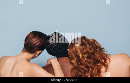 Rearview of three women with different skin tones standing together in a studio. Group of body positive young women embracing their natural bodies. Co Stock Photo