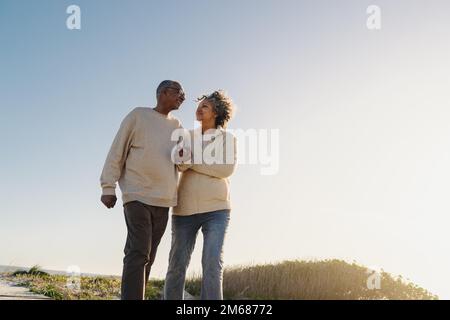 Low angle view of an elderly couple smiling happily while walking down a wooden foot bridge at the beach. Cheerful senior couple enjoying a refreshing Stock Photo