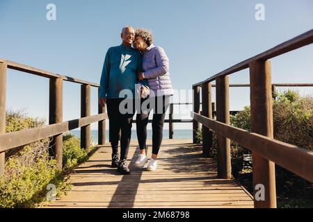 Affectionate senior couple smiling and holding hands while taking a walk along a seaside foot bridge. Romantic elderly couple enjoying a relaxing beac Stock Photo