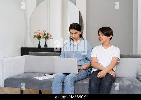 Asian boy sitting near mother using laptop on couch at home,stock image Stock Photo