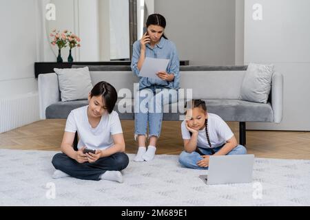 Asian children using devices while mother talking on smartphone and holding paper at home,stock image Stock Photo