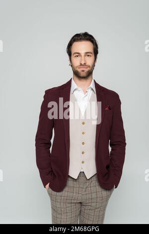 bearded man in vest and red blazer standing with hands in pockets isolated on grey,stock image Stock Photo