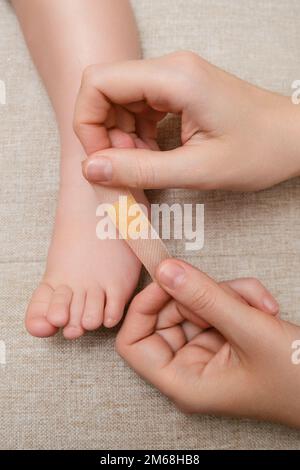 Mother woman sticks a medical band-aid on the toddler baby leg. Mom s hand with sticky wound protection tape and child s foot. Kid aged one year and t Stock Photo