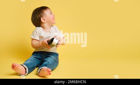 Toddler baby plays with a wireless music speaker on a studio yellow background. Happy child in a white t-shirt and blue jeans listens to music, copy s Stock Photo