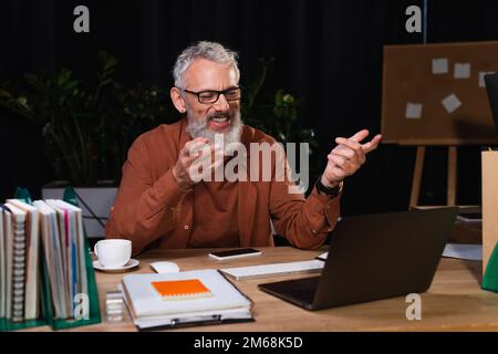 cheerful bearded businessman pointing at laptop while having video call in office at night,stock image Stock Photo