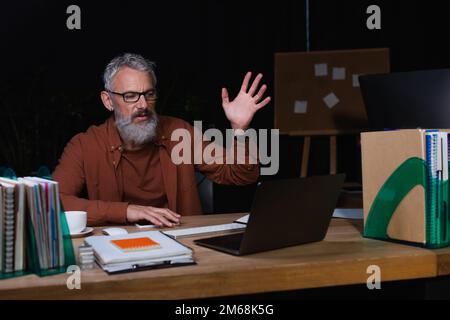 displeased businessman gesturing during video chat on laptop near copybooks in office in evening,stock image Stock Photo