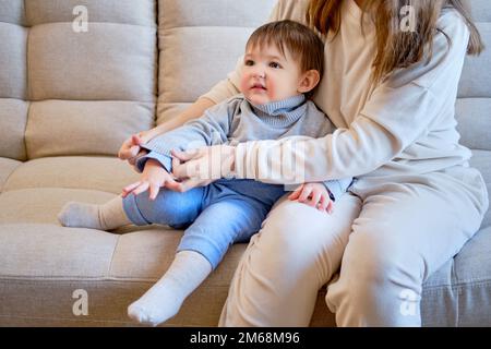 Child is dressed in warm winter clothes for a walk outside. Woman mother puts clothes on toddler baby boy for cold weather. Kid age one year five mont Stock Photo