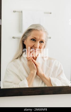 Smiling senior woman looking at her reflection in bathroom mirror Stock Photo