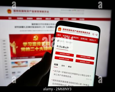 Person holding smartphone with webpage and logo of Chinese state-owned special commission SASAC on screen. Focus on center of phone display. Stock Photo