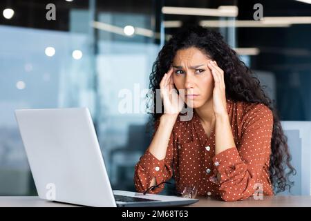 Overtired hispanic business woman working inside modern office, office worker with laptop having severe headache. Stock Photo