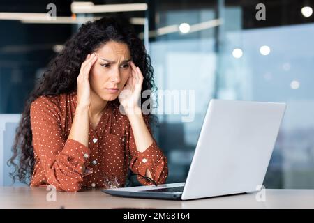 Overtired hispanic business woman working inside modern office, office worker with laptop having severe headache. Stock Photo