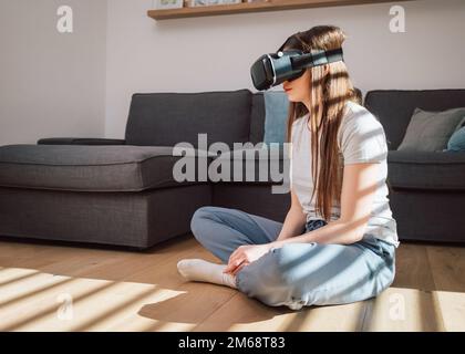Young woman immersing in the virtual world using a VR headset while sitting in the cozy living room Stock Photo