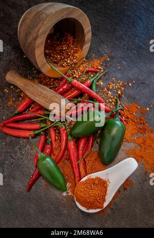 Chilli powder on a white spoon with a wooden pestle in the background surrounded by garlic with red and green chillies on a kitchen bench Stock Photo