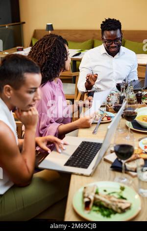 A cheerful group of multiracial colleagues having business lunch in a restaurant and working on the project. Stock Photo