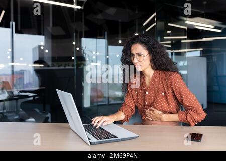 Sick business woman working inside office using laptop while sitting at table, Hispanic woman has severe stomach pain. Stock Photo