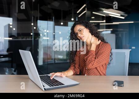 Overtired business woman working inside office with laptop, Hispanic female worker has severe neck pain, massaging muscles with hand. Stock Photo