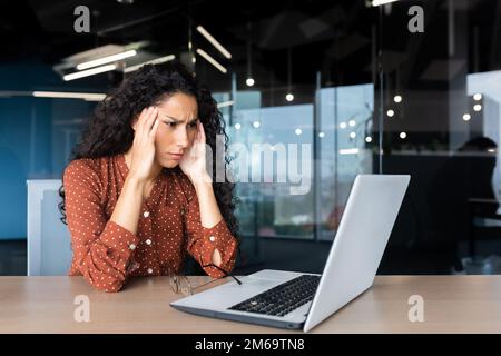 Overtired hispanic business woman working inside modern office, office worker with laptop having severe headache. Stock Photo