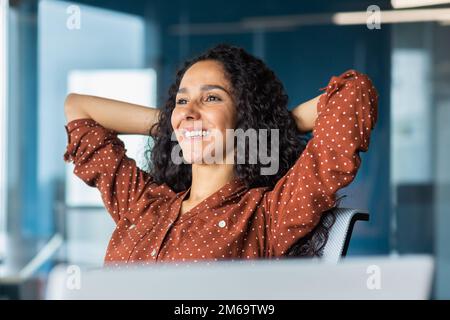 Happy and smiling successful hispanic business woman finished work satisfied with work result and achievement, hands behind head and smiling close up. Stock Photo