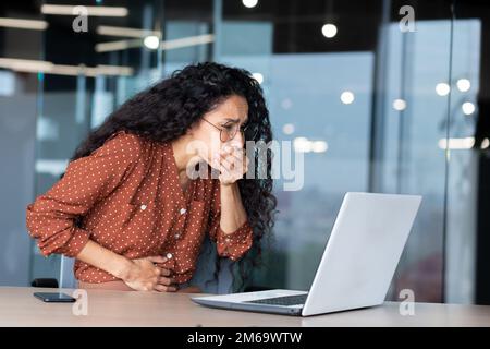 Businesswoman has severe nausea, latin american woman at work sick, working sitting at table with laptop inside office. Stock Photo