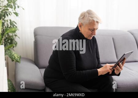 Smiling middle-aged Caucasian woman sit on couch in living room browsing wireless Internet on tablet, happy modern senior female relax on sofa at home Stock Photo