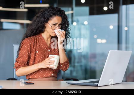 Sick woman working in office, hispanic woman having flu and cold drinking hot tea sneezing and coughing at workplace, using napkins, sitting at table with laptop. Stock Photo
