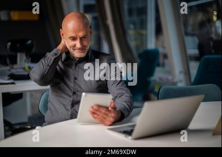 Shot of thinking businessman using digital tablet and laptop while sitting at desk and working in late night at the office. Multitasking. Stock Photo