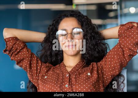 Hispanic business woman with closed eyes dreaming at workplace, close-up female worker with hands behind head thinking serious. Stock Photo
